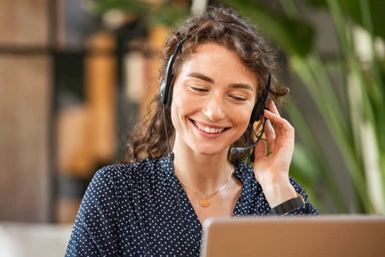 Photo en gros plan d'une jeune femme souriante portant un casque avec micro, en train de travailler devant un ordinateur portable. Elle a des cheveux bouclés et porte une blouse bleue à pois blancs. La scène se déroule dans un environnement intérieur avec des plantes vertes en arrière-plan, donnant une ambiance chaleureuse et professionnelle.