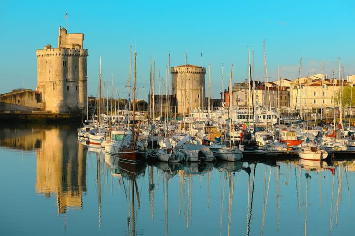 Vue du Vieux Port de La Rochelle, France, avec ses célèbres tours médiévales, la Tour Saint-Nicolas à gauche et la Tour de la Chaîne au centre. De nombreux bateaux sont amarrés dans le port, leurs mâts se reflétant dans l'eau calme. Les bâtiments historiques aux façades claires bordent le port, ajoutant au charme pittoresque de la scène. Le ciel est bleu et dégagé, baignant le port dans une lumière douce et dorée.