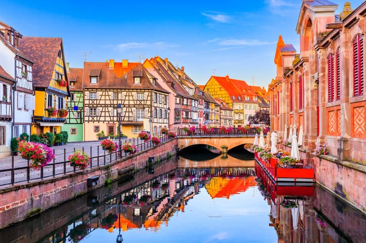Vue pittoresque du quartier de la Petite France à Strasbourg, France. Les maisons à colombages colorées bordent un canal, reflétant leurs façades dans l'eau calme. Des ponts enjambent le canal, décorés de jardinières fleuries. À droite, une terrasse de café avec des parasols blancs s'étend le long du canal. Le ciel est bleu avec quelques nuages, ajoutant une touche vive et ensoleillée à cette scène charmante et typique de l'Alsace.