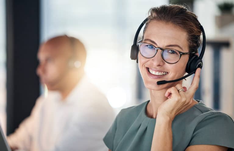 Une femme portant des lunettes et un casque avec microphone sourit et regarde vers la caméra. Elle est vêtue d'un haut vert et travaille dans un environnement de bureau moderne. En arrière-plan, un homme avec un casque similaire est légèrement flou, concentré sur son travail. La lumière naturelle éclaire la scène, créant une ambiance professionnelle et accueillante.