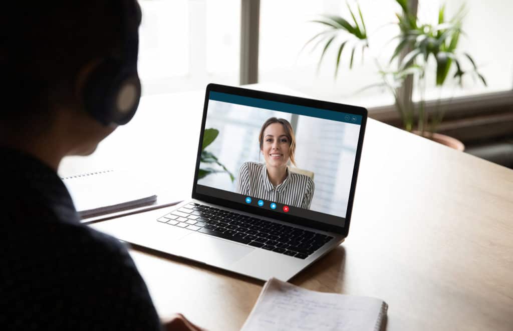 Une personne portant un casque est assise devant un ordinateur portable, participant à une visioconférence. L'écran de l'ordinateur affiche une femme souriante en chemise à rayures, semblant être en pleine conversation. Sur la table, il y a un cahier ouvert et un stylo, tandis qu'en arrière-plan, on aperçoit des plantes vertes près de la fenêtre, créant une ambiance de travail moderne et lumineuse.