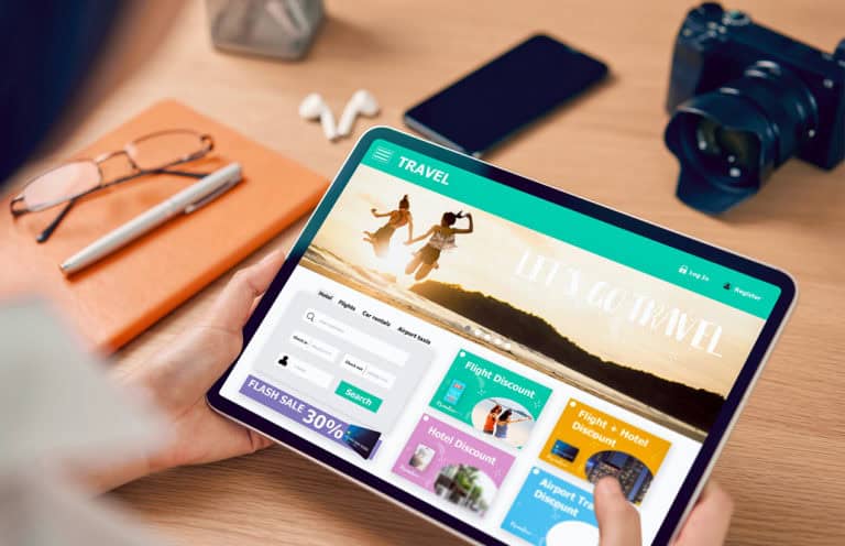 Personne tenant une tablette affichant un site web de voyage avec des offres promotionnelles. L'écran montre des options de recherche pour des hôtels, des vols, des locations de voiture et des taxis d'aéroport, ainsi que des offres de réduction sur les vols et les hôtels. En arrière-plan, sur le bureau, se trouvent une paire de lunettes, un stylo, un carnet orange, un smartphone et un appareil photo.