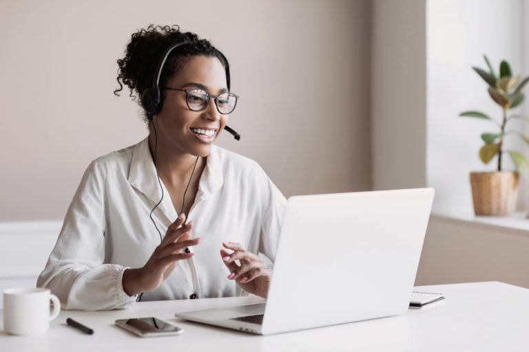 Image d'une femme souriante portant un casque avec micro, travaillant sur un ordinateur portable. Elle est assise à un bureau, avec une tasse, un smartphone et un stylo à côté d'elle. Une plante en pot est visible en arrière-plan près d'une fenêtre, symbolisant l'assistance à distance ou le service client en ligne.