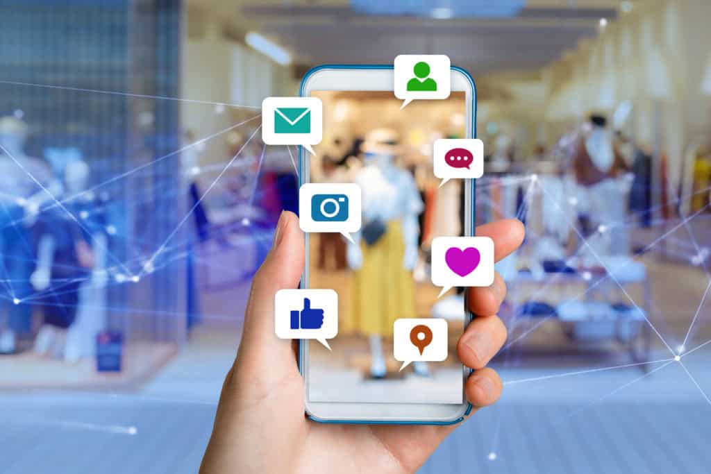 Image d'une main tenant un smartphone, affichant diverses icônes de réseaux sociaux et de communication comme un email, une personne, une bulle de dialogue, un cœur, une caméra, un like et un marqueur de position. L'arrière-plan montre un magasin flou avec des mannequins en vitrine, symbolisant l'intégration des réseaux sociaux dans le commerce et les affaires en ligne.