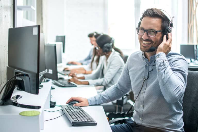 Un centre d'assistance téléphonique avec plusieurs agents assis à leurs postes de travail. Chaque agent porte un casque avec microphone et travaille sur un ordinateur. Au premier plan, un homme souriant, portant des lunettes et une chemise à pois, ajuste son casque tout en regardant son écran. En arrière-plan, d'autres agents sont également engagés dans des appels. L'ambiance est professionnelle et dynamique, illustrant un environnement de support client ou de service après-vente.