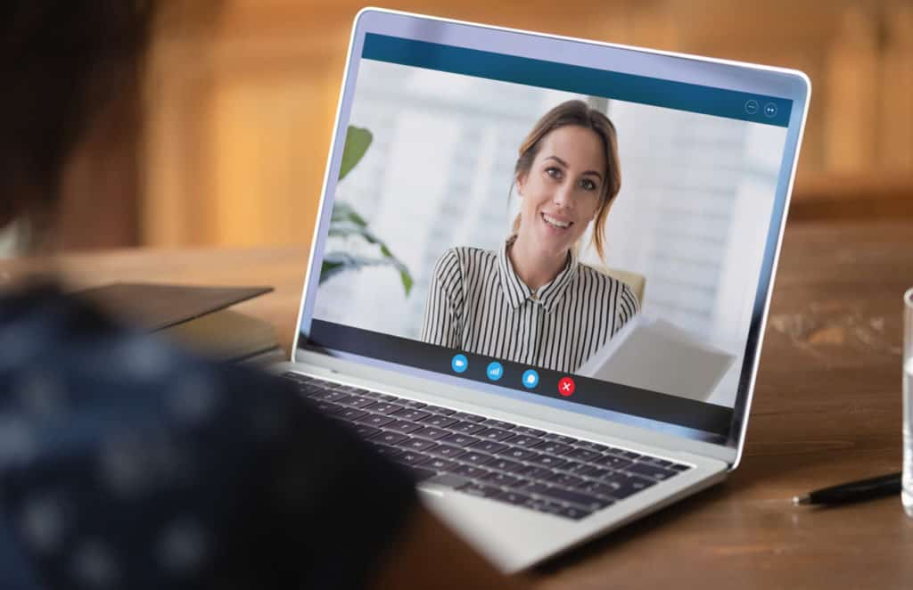 Une personne participant à une vidéoconférence sur un ordinateur portable. L'écran affiche une femme souriante, habillée d'une chemise rayée, regardant vers la caméra. La barre d'outils de l'application de vidéoconférence est visible en bas de l'écran avec des icônes pour la caméra, le micro, la connexion, le chat et la fin de l'appel. En arrière-plan, on peut voir un bureau avec un carnet et un stylo, ainsi qu'un verre d'eau. Cette scène illustre la communication à distance et le travail collaboratif via des outils numériques.