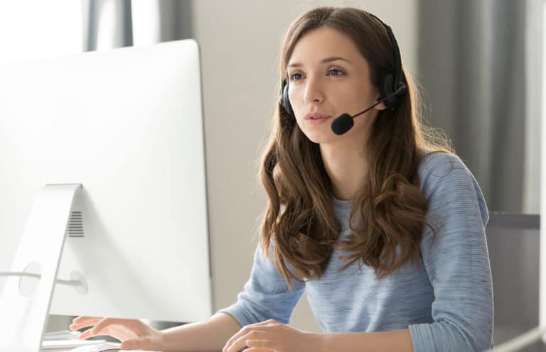 Une jeune femme portant un casque avec micro, assise à un bureau et travaillant sur un ordinateur de bureau. Elle semble engagée dans une conversation ou un appel, probablement offrant un service d'assistance ou de support client. La lumière naturelle éclaire la pièce, créant une ambiance de travail professionnelle et concentrée.