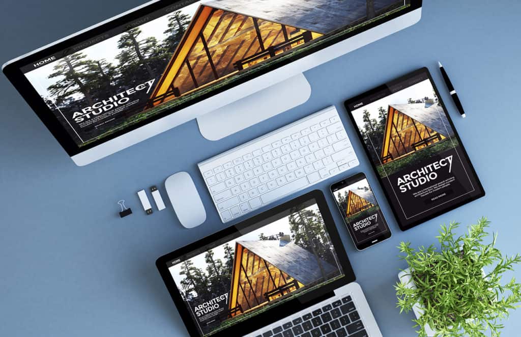 Un bureau avec plusieurs appareils numériques affichant le même site web d'un studio d'architecture, démontrant la conception web réactive. L'écran d'ordinateur de bureau, l'ordinateur portable, la tablette et le smartphone montrent tous l'image d'un bâtiment moderne en bois avec le texte 'Architect Studio'. Sur le bureau, il y a également un clavier sans fil, une souris, des clés USB, une pince à papier, un stylo et une plante verte. Le fond bleu clair de la table ajoute une touche de couleur à cet espace de travail moderne et organisé.