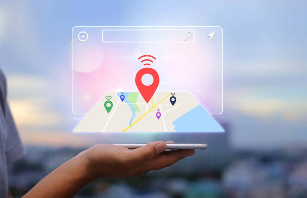 Une main tenant un smartphone d'où émerge une carte virtuelle en trois dimensions avec plusieurs marqueurs de localisation colorés (rouge, vert, bleu et violet). Un grand marqueur rouge est placé au centre, symbolisant une position GPS principale. L'arrière-plan montre un paysage flou de la ville avec un ciel dégradé, évoquant l'utilisation de la technologie de localisation et de navigation sur mobile.