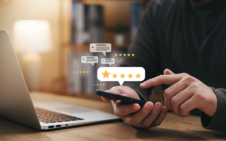 Cette image montre une personne tenant un smartphone, avec des étoiles de notation et des bulles de commentaires flottant au-dessus de l'écran, représentant des avis et des évaluations en ligne. En arrière-plan, un ordinateur portable est visible, ce qui suggère un environnement de travail ou de consultation d'avis via plusieurs appareils. Les étoiles et les commentaires sont des symboles bien connus des plateformes de critique en ligne, où les utilisateurs peuvent évaluer des services ou des produits.