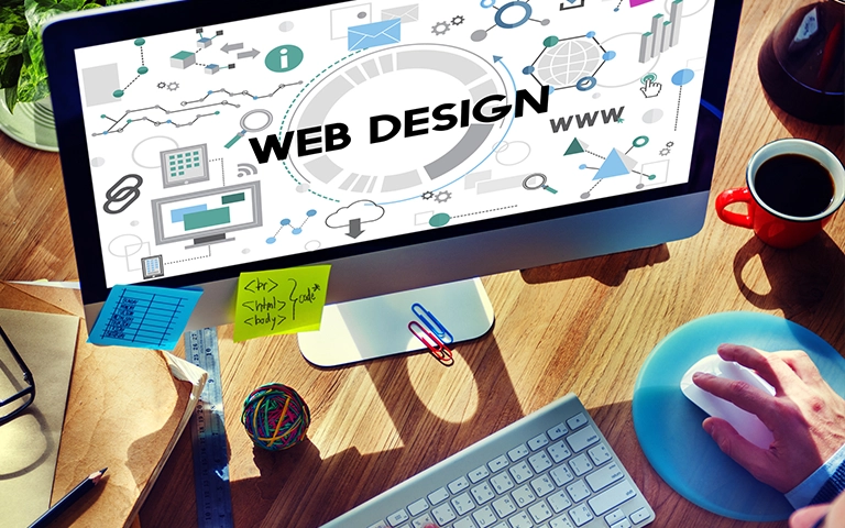 Image d'un bureau avec un ordinateur affichant l'inscription 'Web Design'. L'écran montre des éléments graphiques liés à la conception de sites web, tels que des icônes de graphiques, de réseaux, et de code. Sur le bureau, on trouve des fournitures comme un clavier, une souris, une tasse de café, et des notes autocollantes avec des lignes de code. Une main est visible sur la souris, suggérant une activité de conception ou de programmation.