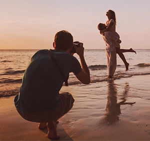 Un photographe en train de prendre une photo d'un couple sur une plage au coucher du soleil. L'homme du couple tient la femme dans ses bras, et ils sont tous deux debout dans l'eau peu profonde, visiblement heureux. La scène est illuminée par une douce lumière dorée provenant du soleil couchant, ce qui crée une atmosphère romantique et paisible.