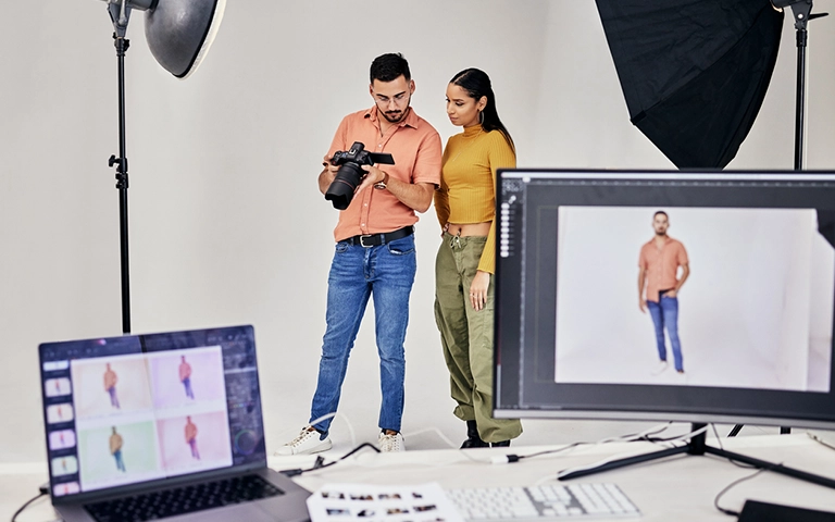 Cette image montre un photographe professionnel en plein travail dans un studio photo, en compagnie d'une cliente ou d'une assistante. Le photographe est en train de vérifier une photo sur son appareil photo numérique, probablement pour s'assurer que le cliché est satisfaisant. En arrière-plan, on peut voir des équipements de photographie tels qu'un grand réflecteur et des lampes de studio. Sur un ordinateur en avant-plan, plusieurs images sont affichées, peut-être des échantillons des prises précédentes. L'atmosphère est professionnelle, avec une attention portée à la qualité et au détail dans la prise de vue.