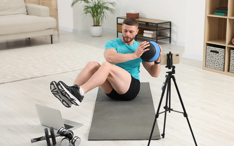 Un coach sportif effectuant un exercice avec un medecine ball sur un tapis de yoga dans une pièce lumineuse. Il est filmé par une caméra montée sur un trépied, tandis qu'un ordinateur portable et des haltères sont posés au sol à côté de lui. La scène illustre une session de coaching en ligne ou un tutoriel d'exercice.