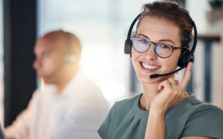 Cette image montre une femme souriante portant un casque avec microphone, ce qui suggère qu'elle fait partie d'une équipe d'assistance clientèle ou de support technique. Elle semble engagée dans une interaction professionnelle, prête à aider les clients ou utilisateurs.