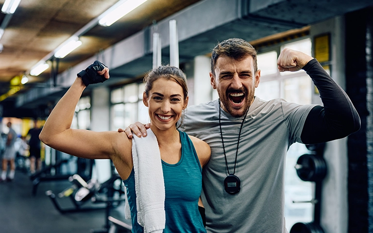 Un coach sportif et une cliente sourient et montrent leurs muscles dans une salle de sport, symbolisant la réussite et la motivation. Le coach porte un chronomètre autour du cou, tandis que la cliente tient une serviette sur son épaule. L'arrière-plan montre des équipements de gym, créant une ambiance dynamique et énergique.